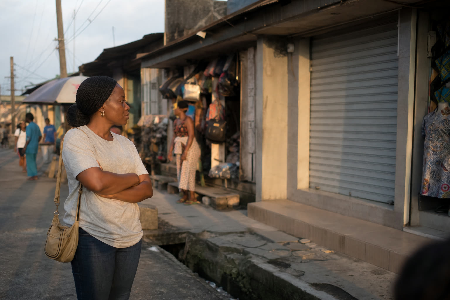 A woman on a street in the early evening, arms folded, looking across at an empty shop with its shutter down.